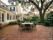 Brick courtyard patio with a round wooden table and four chairs beside a cream building with tall windows and large trees