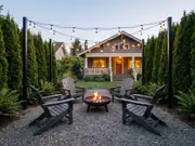 Two Adirondack chairs facing a fire pit under overhead string lights, with a warmly lit house and clipped evergreens behind