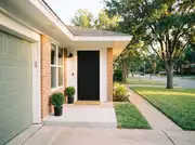 Brick ranch entry with a matte black front door, two boxwood planters, a coir mat and a sage-green garage door