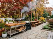 Stainless prep cart with a butcher-block top holds a portable pizza oven and a kettle grill, firewood stacked below