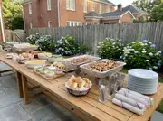 Long teak patio table set as a buffet line with plates, chicken skewers, a taco bar, sides, bread and napkins