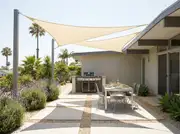 Built-in stucco grill counter under two triangular shade sails, beside a wire-frame dining table on a patio