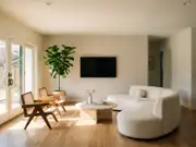 Modern living room with a wall-mounted TV on a white textured wall, beige sofa, round table, and woven chairs by glass doors