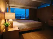 Woven rattan table lamp glowing warmly on a wood nightstand beside a small fern, with blue dusk visible through the window