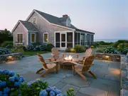Oceanfront stone patio with a fire pit ringed by wooden Adirondack chairs, hydrangeas, and a gray coastal house at dusk