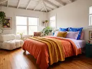White-ceilinged bedroom with a bed in blue, orange and purple bedding, a yellow throw, and a pale couch alongside