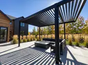 Black aluminum louvered pergola with slats half open, casting striped shadows on a concrete patio