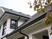Looking up at a white house roofline with black gutters, downspouts, roof vents and window trim in one finish