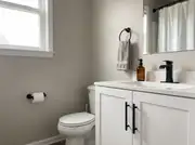 White shaker vanity with matte black bar pulls, faucet, towel ring, and toilet paper holder on greige walls