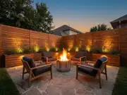 Stone fire pit ringed by four wood lounge chairs with dark cushions, a tall wood privacy fence and shrubs behind at sunset