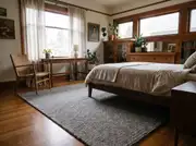 Large grey wool-look area rug under a wood bed frame, with a woven armchair's front legs resting on the rug near a desk