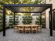 Paved courtyard dining area with a wooden table and chairs under a black pergola and vine-covered brick-and-white walls