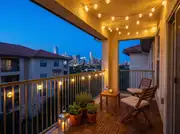 Warm globe string lights looped across a stucco balcony ceiling and railing at dusk above a small teak table