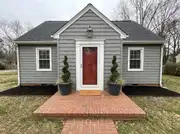 Gray shingled cottage with a red front door flanked by two charcoal planters holding spiral boxwoods