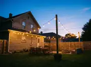 Backyard at dusk with string lights spanning from the house eave to cedar posts above a dining table and fire pit