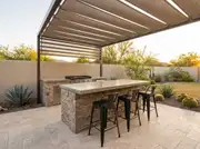 Stone-clad grill island with a concrete bar top and three black stools under a louvered pergola in a desert backyard