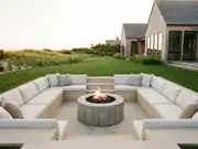 Curved white sectional around a round concrete gas fire pit with flames on a stone patio, a row of modern houses behind