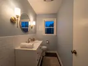 Brass wall sconces glowing on both sides of a round mirror above a white vanity at dusk, subway tile below