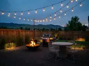 Gas grill and a low steel fire bowl on a gravel patio at dusk, string lights strung above a round table with wire chairs