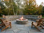 Circular stone fire pit with flames inside a low curved stone seat wall, two wood rocking chairs and an autumn tree behind