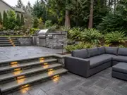 Tiered stone patio with step lights, a gray outdoor sectional along the upper terrace, and a landscaped yard of tall trees