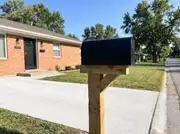New matte black mailbox on a straight wooden post beside a clean concrete driveway and a brick ranch