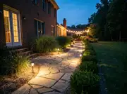 Flagstone path beside a brick house lit by small warm solar path lights leading to a string-lit patio