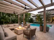 Beige sectional around a square fire pit under a wood pergola with black ceiling fans, potted plants and a pool by the house