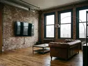 Industrial loft living room with a wall-mounted TV on an exposed brick wall, a dark leather tufted sofa, and city windows