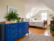 Bedroom with a slanted ceiling, striped wallpaper, and a glossy blue dresser topped with a fern and framed art