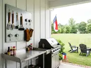 Galvanized pegboard holding grill tools, mitts and thermometers beside a gas grill on a covered porch