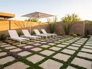 Modern patio with pavers set in purple and green ground cover, white lounge chairs under a large metal shade sail at sunset