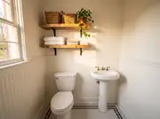 Two wood shelves on black brackets above a toilet, holding folded towels, seagrass baskets, and a pothos