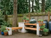 DIY stacked cinder-block counter with wood plank shelves holding a green kamado grill and potted herbs against a wood fence