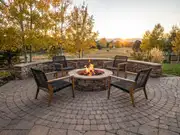Lit stone fire pit ringed by four wood-and-woven chairs on a curved brick patio, with autumn trees at sunset
