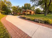 A concrete driveway and walkway edged with a brick paver border leading to a brick ranch home