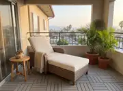 All-weather wicker lounge chair with a cream cushion and throw on gray deck tiles beside two potted palms