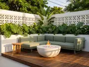 Green L-shaped sofa and a flaming round fire pit on a tiled deck with a wood side table, tropical plants and a white wall