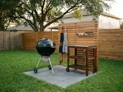 Freestanding black kettle grill on a small paver pad beside a slatted wood prep cart with hanging aprons and tongs