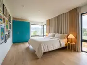 Modern bedroom with a neatly made bed, beige curtains, framed art, and a teal wardrobe against windows onto green hills