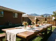 Outdoor dining table with a linen runner, ceramic plates, mason-jar flowers, a fruit bowl and balloons on the fence