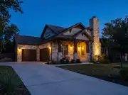 Stone-fronted house at dusk with porch sconces, garage lanterns and a chimney uplight all glowing warm amber