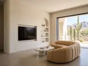 Modern living room with a TV recessed into a wall of built-in shelves, a curved tan sectional, and desert-view windows