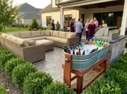 Wraparound wicker sectional and a galvanized drink tub on stands, guests gathered near a stone-clad grill counter on a patio