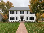 Symmetrical white colonial with black shutters on every window and a brick walkway through an autumn lawn