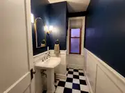 Tiny half bath with deep navy walls, white wainscoting, a brass arched mirror, and a checkerboard floor