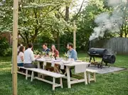 Group seated at a long table under string lights while a black barrel smoker gives off smoke on a grassy backyard