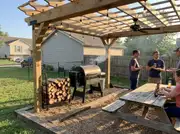 Wood pergola with market lights over a pellet smoker and picnic table, guests standing nearby on a gravel pad