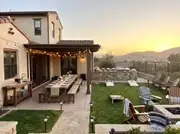 Spanish-style home's backyard at sunset with a string-lit dining table, drink station, buffet, cornhole and fire pit
