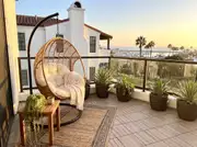 Rattan hanging egg chair on a black stand with a jute rug, side table and potted agaves overlooking a harbor